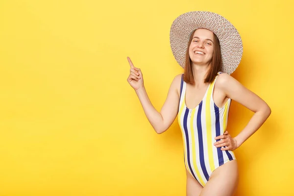 縞模様の水着と帽子を身に着けている満足の女性は 空の領域を指して黄色の背景に隔離され 広告のためのスペース — ストック写真