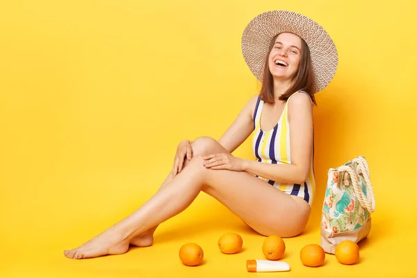 面白いです幸せな女性身に着けていますストライプ水着と帽子隔離上の黄色の背景座って笑います楽しい時間で夏のリゾート — ストック写真