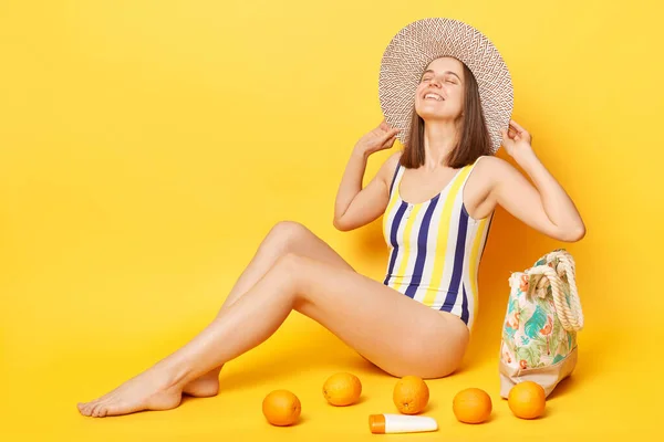 喜びに満ちた女性は縞模様の水着と帽子を身に着けています幸せを表現するビーチで黄色の背景日光浴に隔離されました — ストック写真