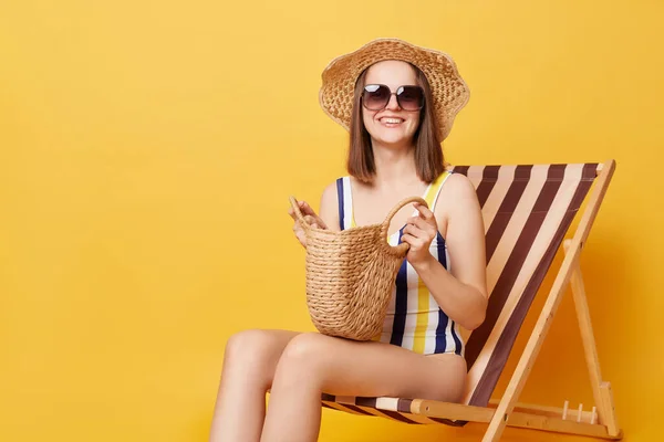 縞模様の水着と帽子を身に着けている若い女性は 黄色の背景の上に隔離されたデッキチェアに座っていますリゾートでビーチバッグ日光浴を保持 — ストック写真