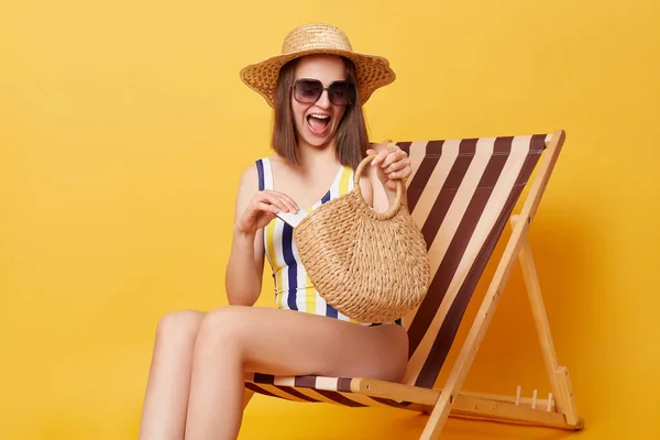 快乐的年轻女子坐在甲板椅子上 与黄色背景隔离 提着旅行袋和防晒霜 欢度她的假日日光浴 — 图库照片