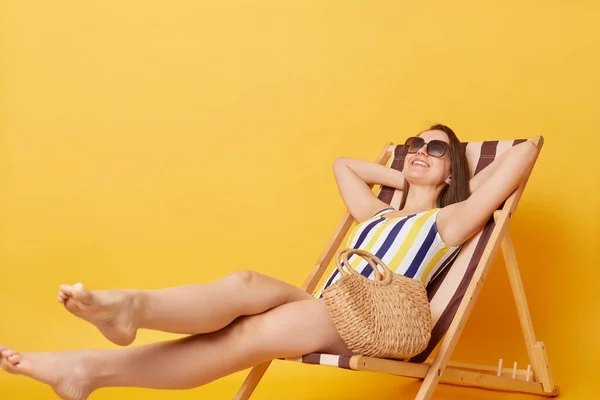 リラックスした満足度の高い女性を身に着けていますストライプワンピース水着はデッキチェアに座って黄色の背景に隔離されています日光浴オンリゾート良好な夏休みを持っています — ストック写真