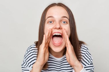 Kahverengi saçlı, çizgili tişörtlü, mutlu bir kadın. Gri arka planda tek başına duruyor. Ağızdan çığlık çığlığa bağırıyor..