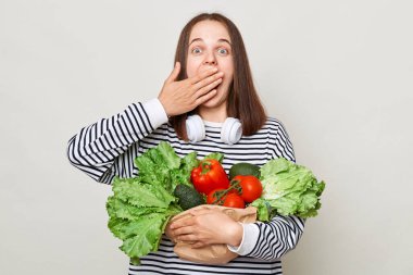 Kahverengi saçlı, çizgili tişörtlü, gri arka planda dikilip yüksek fiyatlı ideal beslenme için mevsimlik sebze alan sürpriz bir kadın..