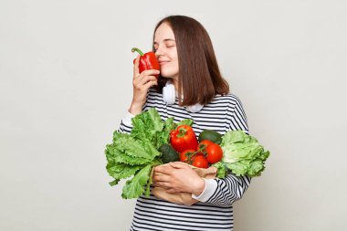 Sağlıklı, kahverengi saçlı, çizgili tişörtlü, gri arka planda tek başına duran sağlıklı bir kadın beslenme düzenini koruyor. Elinde taze biber kokusu var..