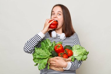 Kahverengi saçlı, çizgili tişörtlü, gri arka planda tek başına duran aç bir kadın elinde taze mevsimlik sebzelerle çan biberi ısırıyor..