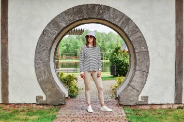 Çizgili tişört giymiş, panama giymiş, parkın girişinde poz veren, sokaklarda gezen gülümseyen kadın portresi..