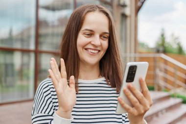 Neşeli, esmer, çizgili tişört giyen, video mesajı alan, el sallayan, el sallayan büyük pencereli binanın yanında poz veren neşeli, esmer bir kadın..