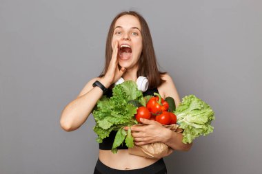 Gri arka planda saklanmış organik sebzeler taşıyan heyecanlı beyaz kadın, el ele bağırarak piyasada indirimlerle ilgili duyurular yapıyor..