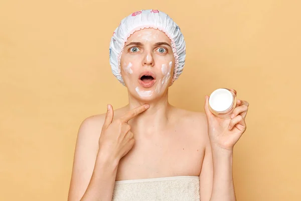 Face peeling, skin care. Portrait of shocked surprised woman applies moisturising facial mask, gets good care, puts nutritious cream, looks at camera with open mouth wears shower cap.