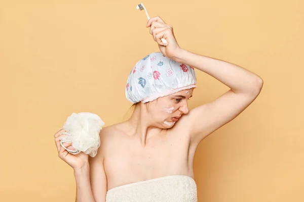 Caucasian young woman disgusted by the scents of her armpits standing against beige wall wearing shower cap and wrapped in towel holding sponge and toothbrush