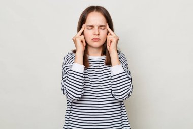 Üzgün, sağlıksız, kahverengi saçlı, çizgili gömlek giyen, gri arka planda yalnız poz veren, baş ağrısı çeken, şakaklarını kapalı gözlerle ovuşturan bir kadın..