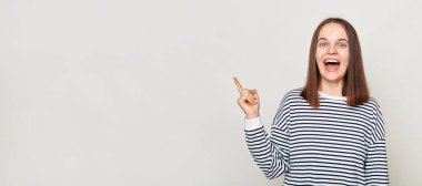 Çekici, kahverengi saçlı, çizgili tişört giyen, gri arka planda yalnız poz veren, reklam alanına bakan, mutlu yüzlü, neşeli bir kadın..