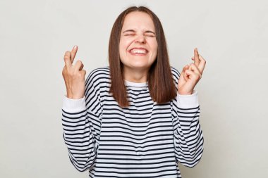 Making wish. Praying for good luck. Smiling hopeful brown haired woman wearing striped shirt posing isolated over gray background standing with crossed fingers and closed eyes. clipart