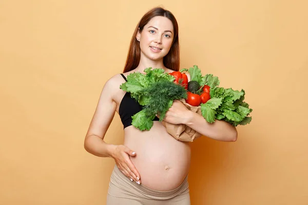 暗い髪の魅力的な妊娠中の女性は彼女の裸のおなかを示すベージュの背景に孤立ポーズ新鮮な野菜を包含する彼女の裸のおなか妊娠中の有機食品 — ストック写真