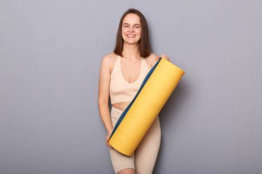 Çekici güzel sporcu kadın bej bluz giyiyor ve yoga minderi tutan taytlar gri arka planda izole edilmiş fiziksel egzersizin keyfini çıkarıyorlar..