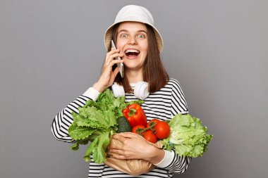 Gri arka planda sebze tutan heyecanlı kadın akıllı telefondan konuşuyor ve heyecanlı bir ifadeyle kameraya bakıyor..