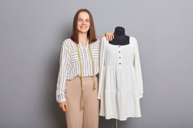 Tekstil üretim süreci. Başarılı bir dikiş işi. Profesyonel moda tasarımcısı. Yeni elbisenin yanında duran güzel, mutlu kadın lağımı gri arka planda izole edilmiş..