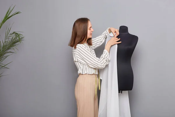 Custom clothing boutique. Small clothing workshop. Side view of smiling hardworking woman tailor standing near new dress on mannequin iisolated over gray background.