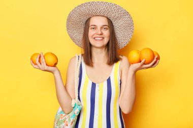 Tek parça çizgili mayo ve hasır şapka giyen gülümseyen neşeli kadın sarı arka planda izole bir şekilde duruyor ve tatilde dinlenen oraganik meyvelerle duruyor..