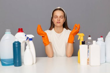 Gri arka planda izole edilmiş temizlik deterjanları ile işyerinde poz veren rahat, yorgun bir kadın. Kapalı gözlerle yoga yapıyor, sıkı çalıştıktan sonra sakinleşmeye çalışıyor..