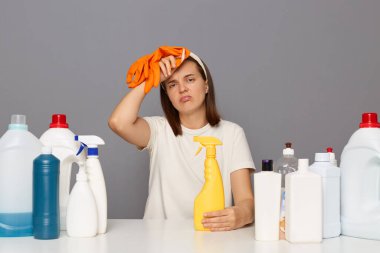 İş yerinde çamaşır deterjanıyla poz veren yorgun bir kadın gri arka planda izole edilmiş uzun saatler çalışıyor yorgun bir ifadeyle kameraya bakarken baş ağrısı çekiyor..