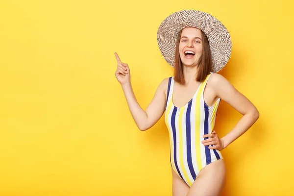 あなたのテキストのために広告スペースを指して笑う黄色の背景の上に隔離された縞模様のワンピース水着と藁帽子を身に着けている喜びの女性 — ストック写真