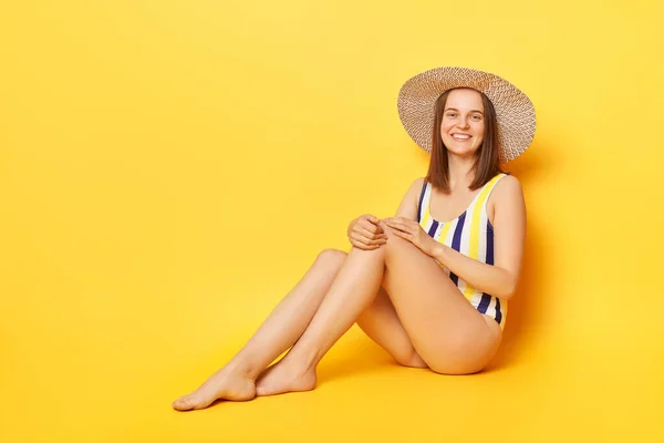 笑顔美しいです若いです女性身に着けていますストライプワンピース水着と麦わら帽子ポーズ孤立した上に黄色の背景見ます上のカメラリラックス夏のリゾート — ストック写真