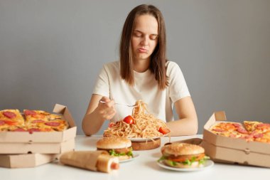 Kahverengi saçlı, beyaz tişörtlü, gri arka planda izole edilmiş fast food 'un arasında oturan canı sıkkın kadın çatal yemeyi sevmiyor..