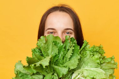 Kilo verme hedefleri. Zayıflatıcı yemek planı. Yeşil ve besleyici. Yüzünün yarısını marul yeşili sebzelerle kaplayan neşeli kadın sarı arka planda izole edilmiş kameraya bakıyor..