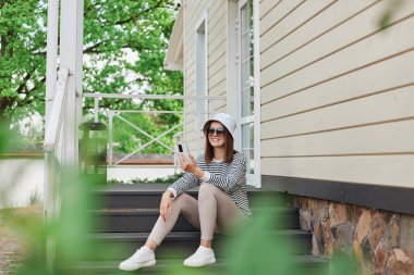 Çizgili gömlek ve panama giyen pozitif hipster kadın ev verandasında oturup internet mesajları okuyarak internete kaymayı seviyor..