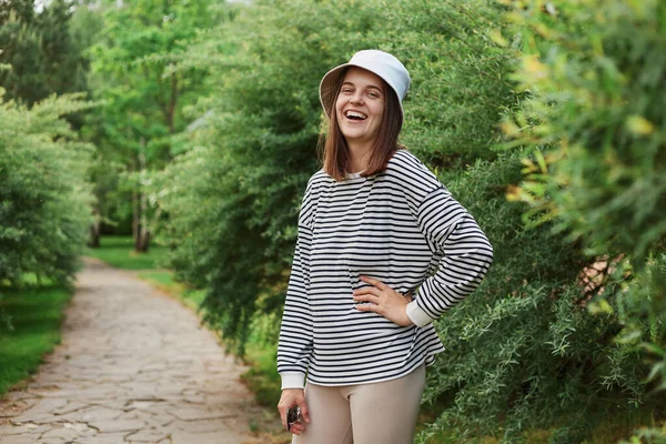 一位黑头发的年轻女子站在绿叶旁笑着 矮树丛手挽手 臀部穿着衬衫 全神贯注地看着她 — 图库照片