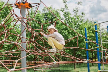 Üzüntülü küçük bir kız, günlük giysiler ve beyzbol şapkası takıyor, yeşil parktaki oyun parkında oynuyor, kötü bir ruh halinde, negatif duygularını ifade ediyor, yalnız başına sıkılmış hissediyor..
