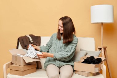 Koyu renk saçlı tatmin olmuş bir kadın bej rengi duvarları olan kutular arasında koltukta oturuyor. Dükkandan sipariş alıyor, paketleri yeni kıyafetlerle açıyor..