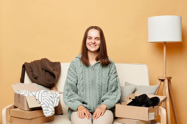 幸せな満足女性座っていますソファの上に箱の中に服を着ていますベージュの壁に対して新しいアパートに移転彼女の持ち物をアンパック彼女の笑顔カメラ — ストック写真