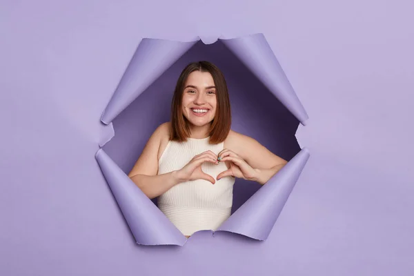 Smiling cheerful brunette woman breaking through purple paper hole wearing casual white top showing heart gesture with hands looking at camera with happy face.