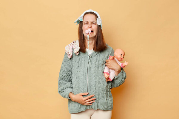Crying sad unhappy young pregnant woman wearing warm knitted sweater with baby clothing isolated over beige background expressing negative emotions touching her belly.