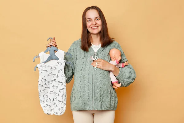 喜びます満足若いです妊娠中の女性とともに赤ちゃんの体のスーツは ベージュの背景に隔離された人形を保持しています子供の服を示すカメラを待っている子供の妊娠を楽しむ — ストック写真