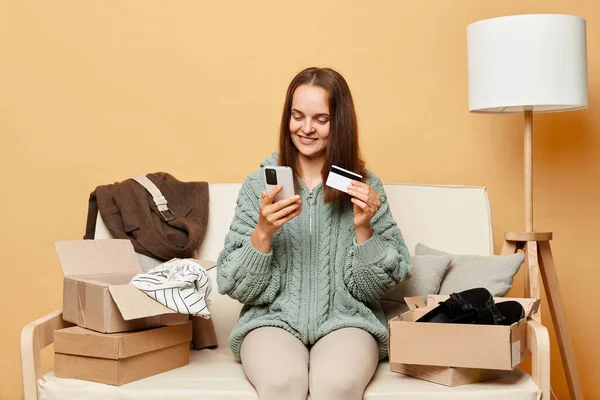 Attractive delighted woman sitting on sofa among boxes with clothing against beige wall ordering clothing in internet store holding smartphone and credit card entering data for paying.