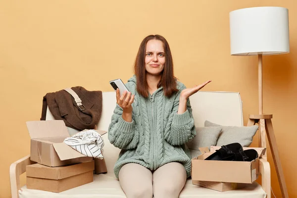 stock image Unhappy sad woman sitting on sofa among boxes with clothing against beige wall using mobile phone with upset expression get wrong parcel from internet shop.