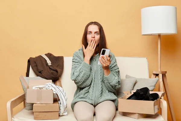 携帯電話を使用してカートンボックスの間でソファに座ってニットセーターを着てショックを受けた不快感の女性は 彼女が間違った注文をオンラインショッピングを行う実現 — ストック写真