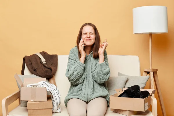 Stock image Hopeful woman sitting on sofa among carton box talking on mobile phone crossing fingers making wish praying for good luck for changing her order.