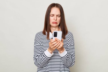 Üzgün, kahverengi saçlı, çizgili tişört giyen, gri arka planda yalnız başına duran, mutsuz yüzlü, hüzünlü bir mesaj okuyan yetişkin kadın..