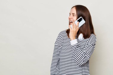 Heyecanlı kahverengi saçlı, çizgili tişört giyen, gri arka planda izole bir şekilde duran, cep telefonuyla konuşan ve reklam için fotokopi odasına bakan yetişkin bir kadın..