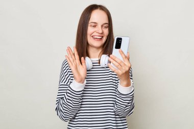 Neşeli, kahverengi saçlı, çizgili tişört giyen, gri arka planda tek başına duran ve elinde akıllı bir telefonla el sallayan bir video olan yetişkin kadın..