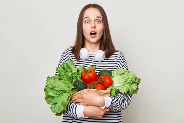 Şok olmuş kahverengi saçlı genç kadın, gri arka planda izole edilmiş çizgili günlük gömleği olan taze sebze buketini kucaklıyor ve büyük gözleriyle kameraya bakıyor..