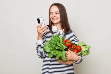 Neşeli kahverengi saçlı genç bir kadın, gri arka planda izole edilmiş çizgili günlük gömleği giyerek bir buket taze sebzeyi kucaklıyor. Cep telefonu faturalarını internetten buluyor..