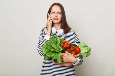 Üzgün, hayal kırıklığına uğramış, kahverengi saçlı genç bir kadın bir buket taze sebzeyi kucaklıyor, çizgili, sıradan bir gömlek giyiyor ve gri arka planda cep telefonuyla konuşuyor.