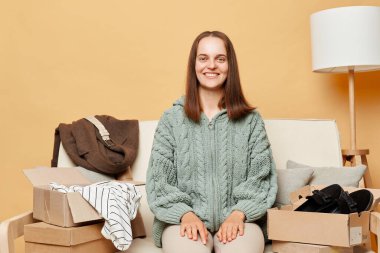 Gülümseyen mutlu kadın, örülmüş süveter ve sipariş edilmiş giysiler içinde koltukta bej duvarın karşısında oturmuş online alışverişin keyfini çıkarıyordu..