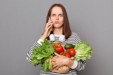 Üzgün, mutsuz bir kadın akıllı telefondan konuşuyor, taze sebzeleri kucaklıyor, gri arka planda izole edilmiş çizgili tişört giyerek marketten dönüyor, şaşkın bakışlarla kameraya bakıyor..